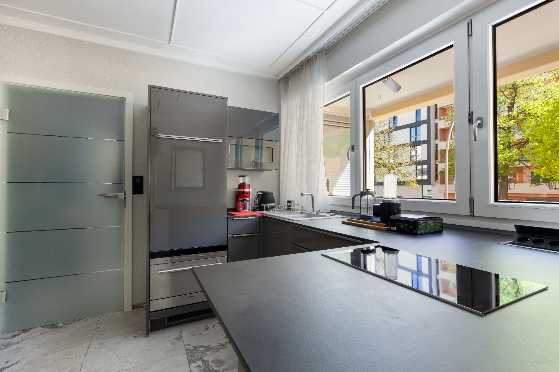 Moderne Küche mit grauen Schränken, großem Fenster und Glastür, Blick auf Wohngebäude draußen.