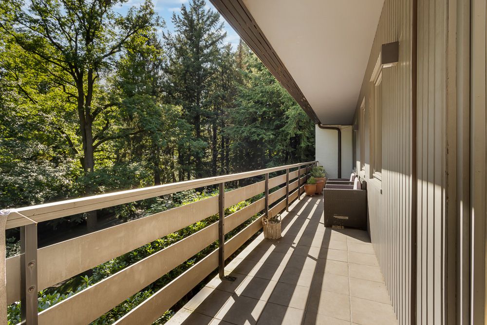 Langer Balkon mit Geländer, Fliesenboden und Blick auf grüne Bäume, Gartenmöbel und Pflanztöpfe sichtbar.