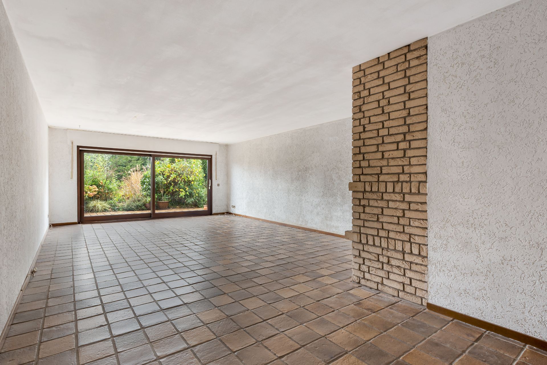 Leeres Wohnzimmer mit Fliesenboden, Sichtmauerwerk und großen Fenstern zum Garten.