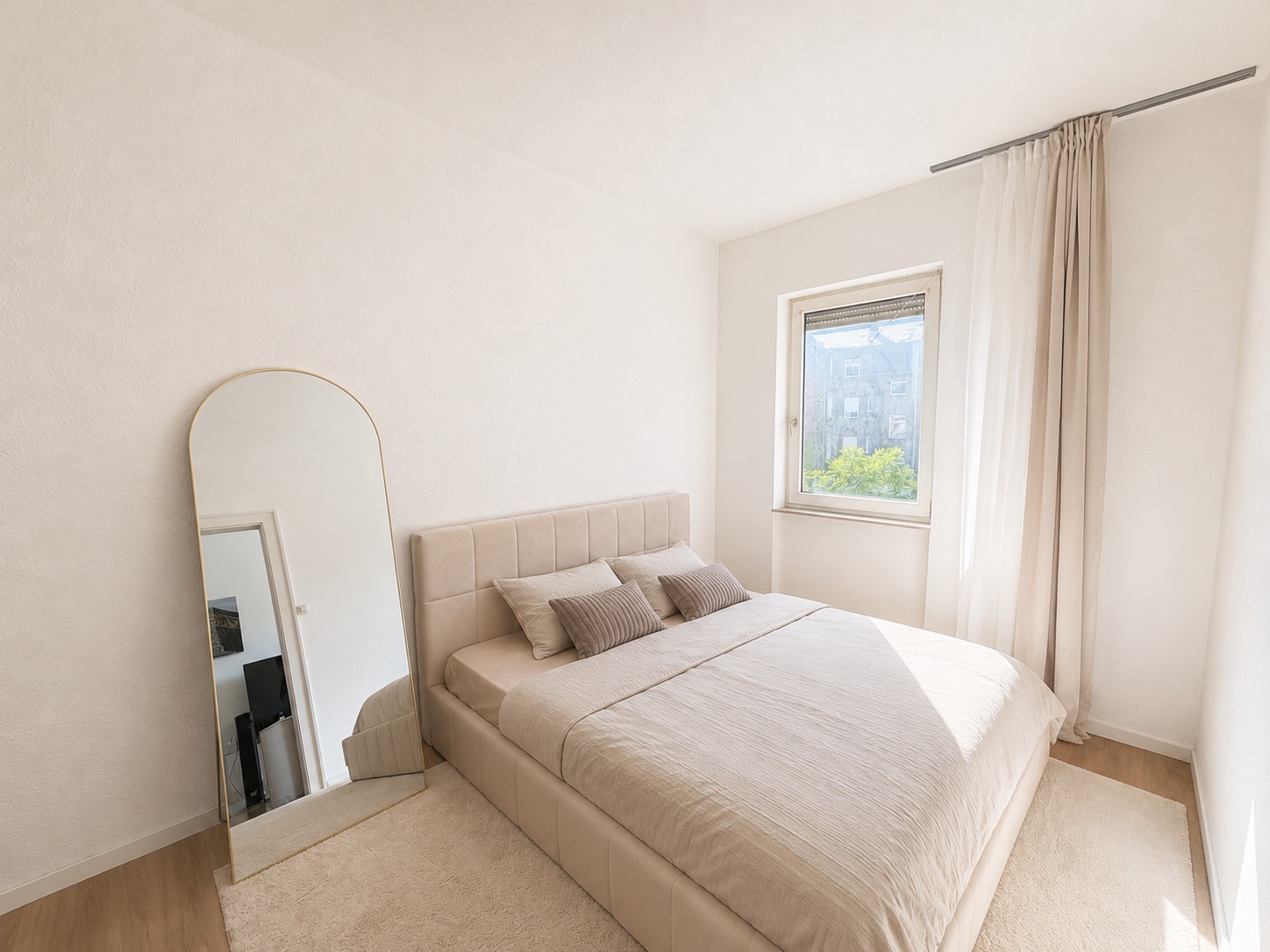 Helles Schlafzimmer mit Doppelbett, großem Spiegel und Fenster mit Vorhang.