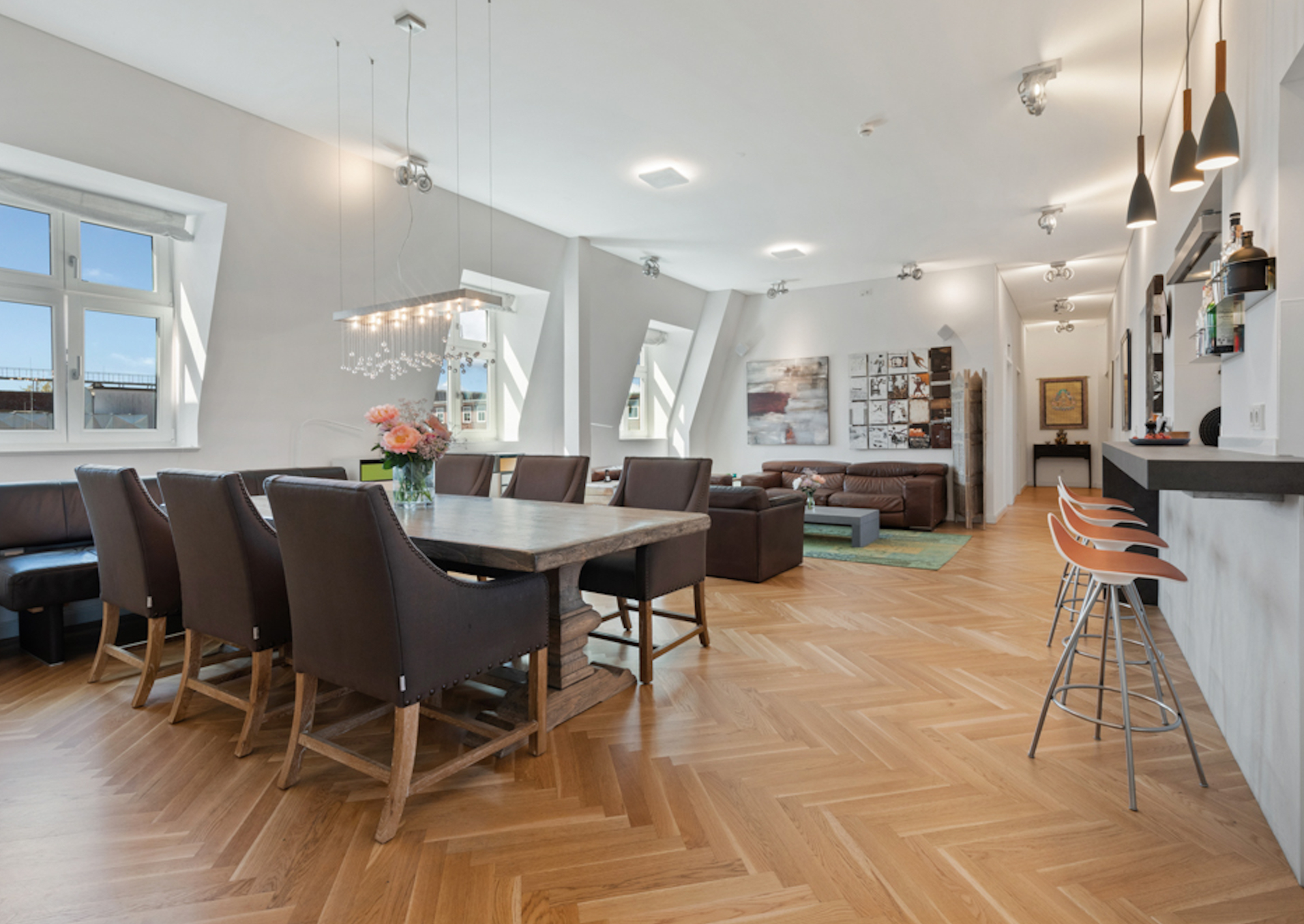Helles Wohnzimmer mit Esstisch, braunen Stühlen, Sofa, Barhockern und Holzboden.