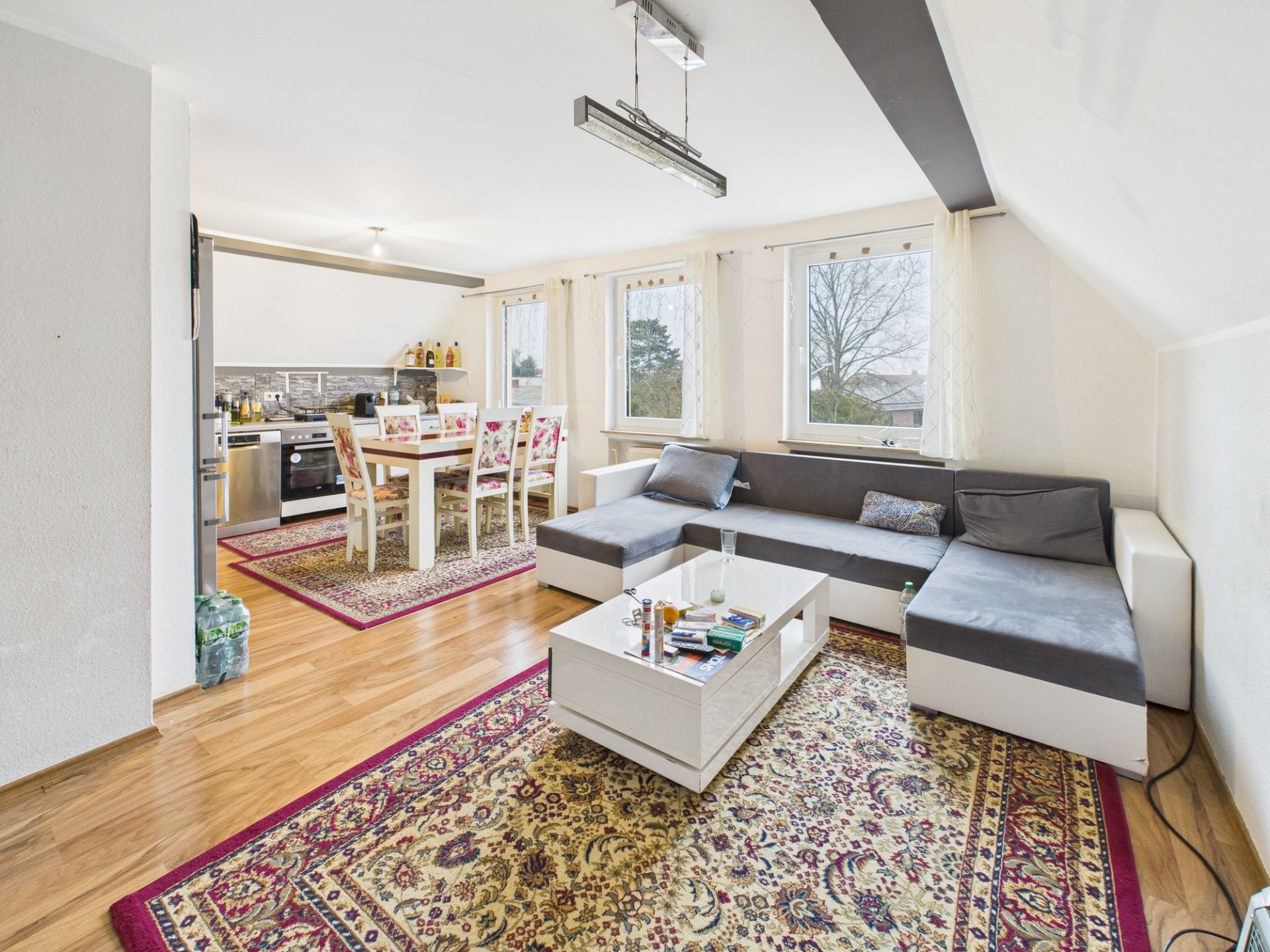 Helles Wohnzimmer mit Ecksofa, Couchtisch, Esstisch und offener Küche, Holzboden und Teppichen.