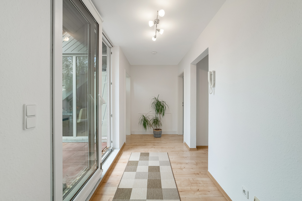 Heller Flur mit Holzboden, großem Fenster, Teppich und einer Topfpflanze am Ende.