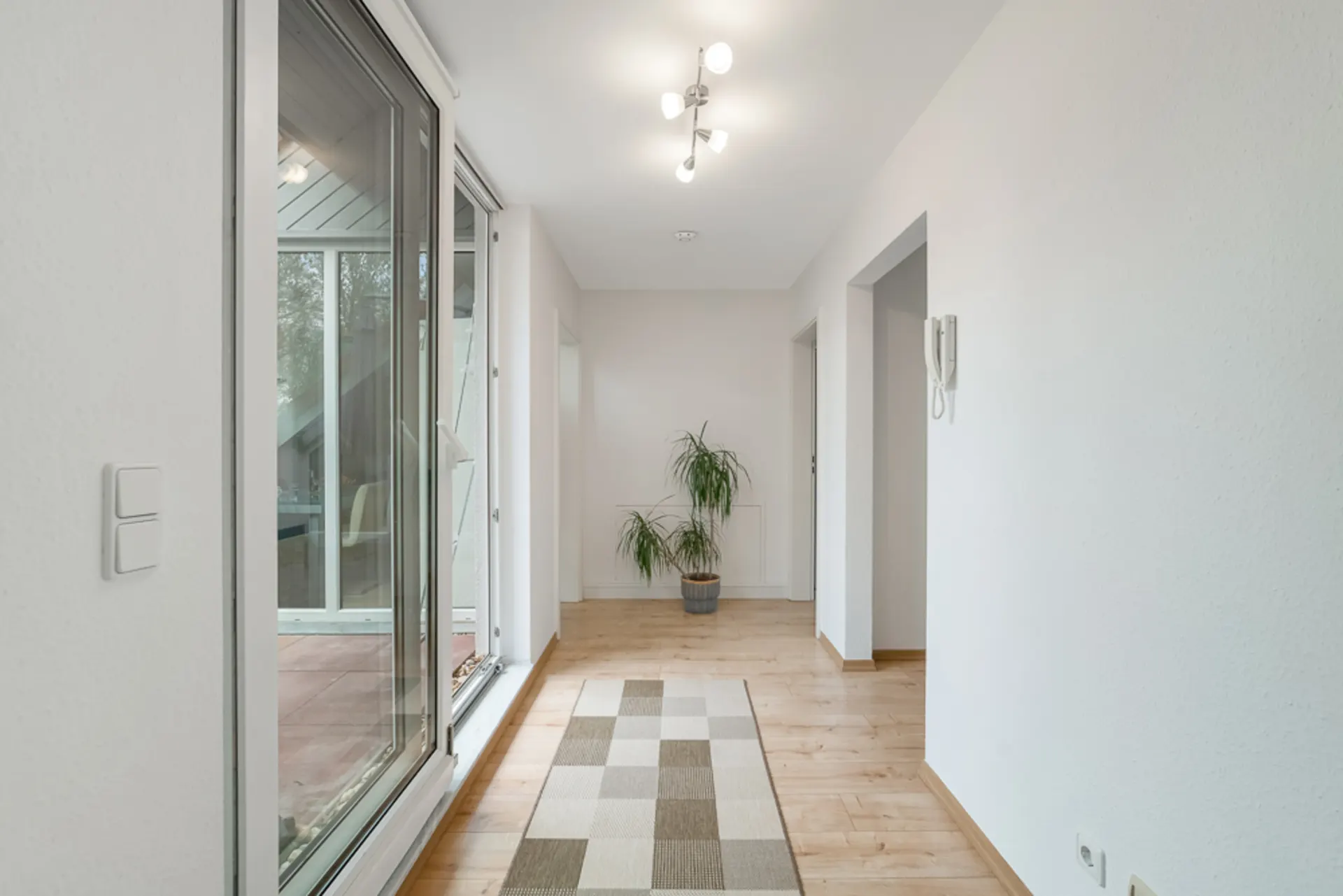 Heller Flur mit Holzboden, großem Fenster, Teppich und einer Topfpflanze am Ende.