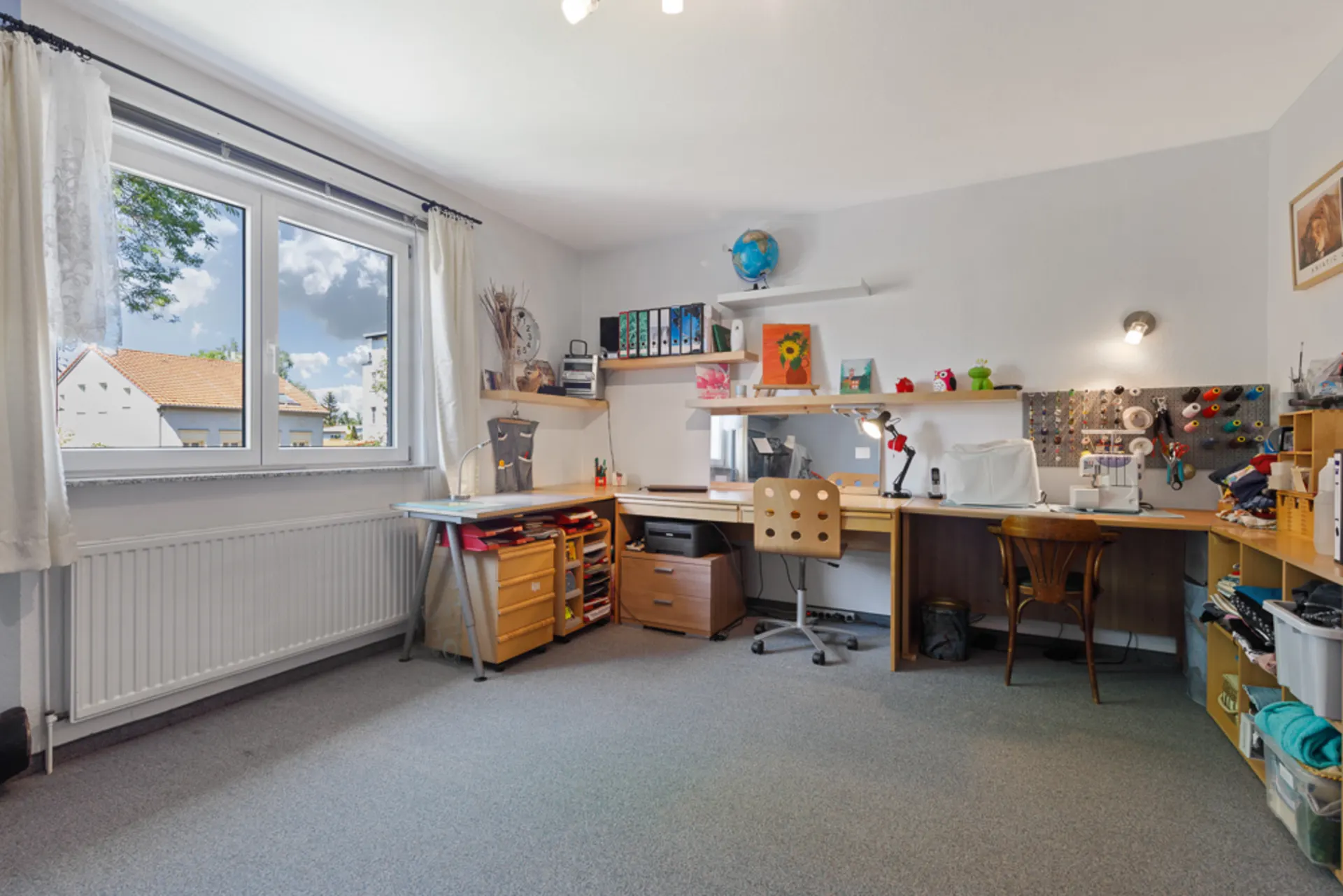 Helles Arbeitszimmer mit großem Fenster, Schreibtischen, Regalen und Nähutensilien.