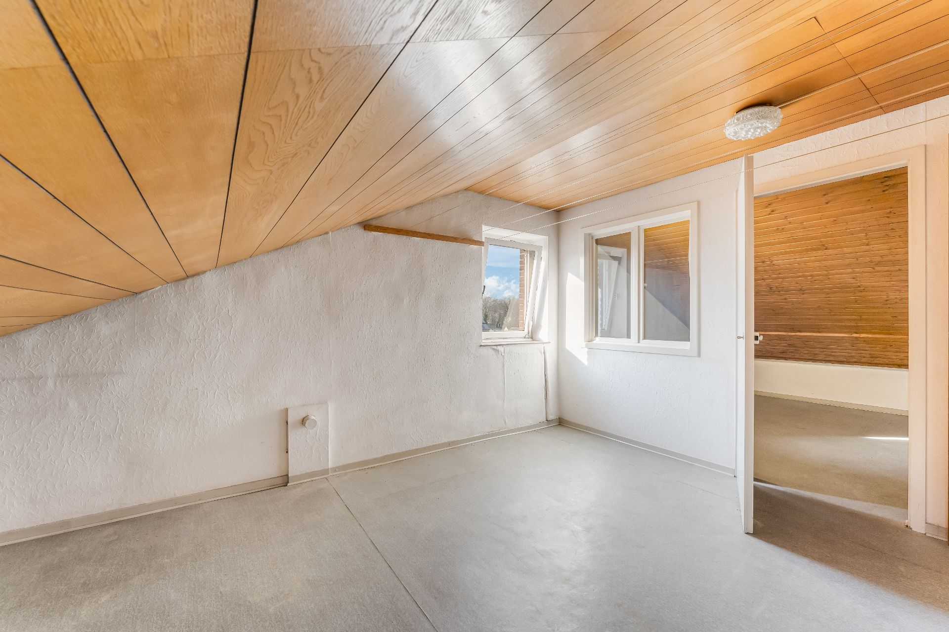 Heller Raum mit Holzdecke, Dachschräge, Fenster und offener Tür zu weiterem Zimmer.