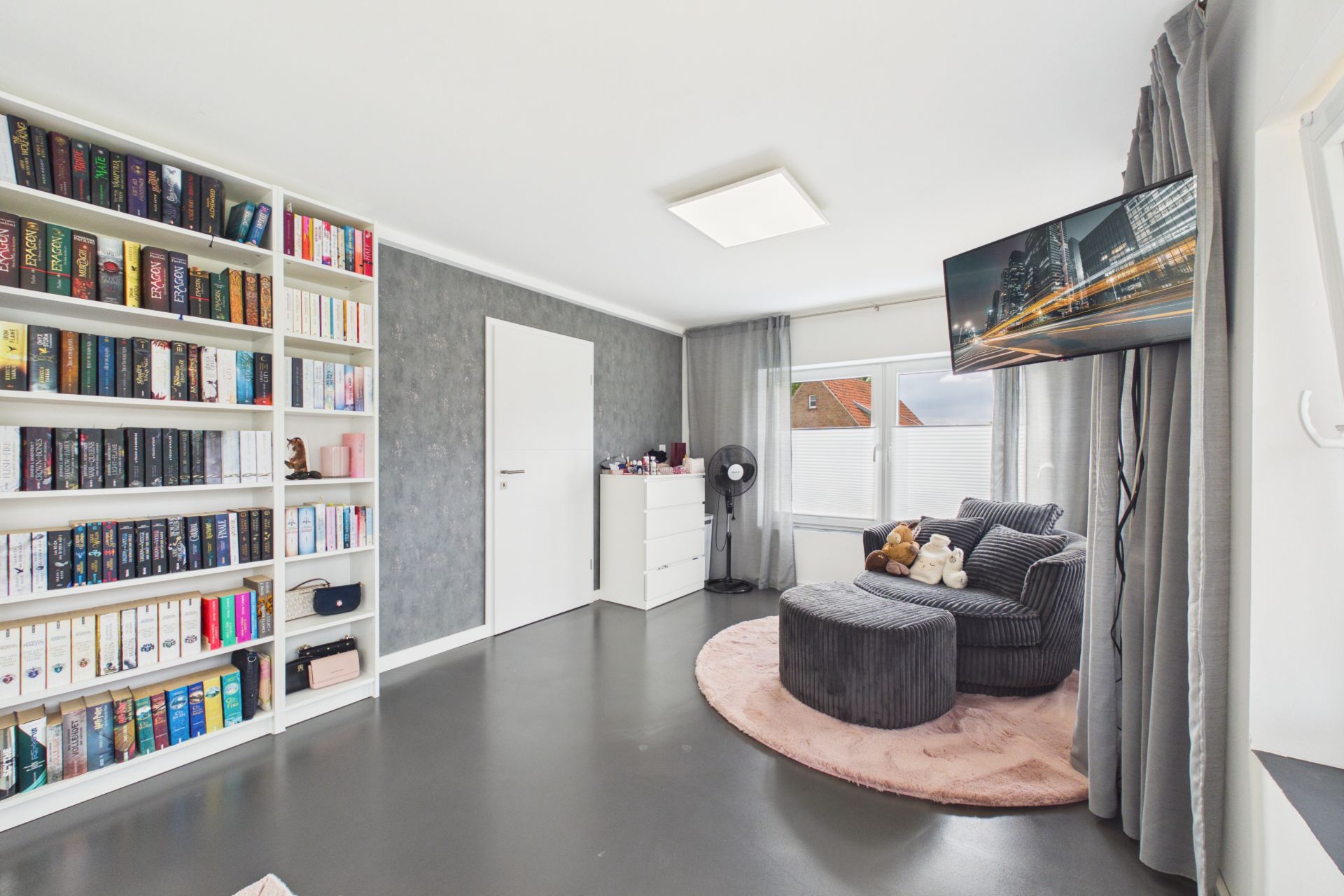 Helles Zimmer mit Bücherregal, Sessel, Fernseher und großem Fenster mit Vorhängen.