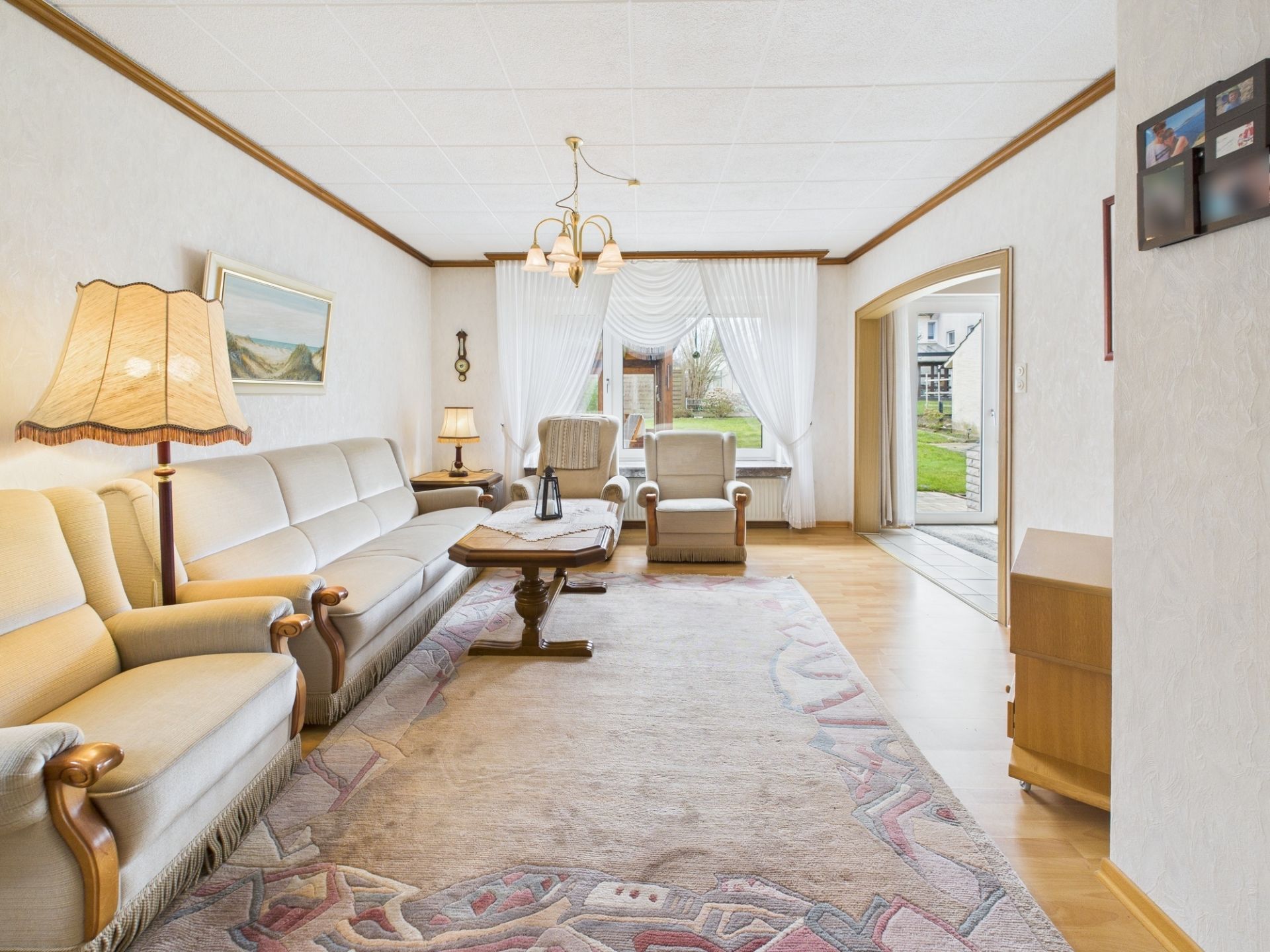 Helles Wohnzimmer mit Sofa, zwei Sesseln, Teppich und großem Fenster mit Blick nach draußen.