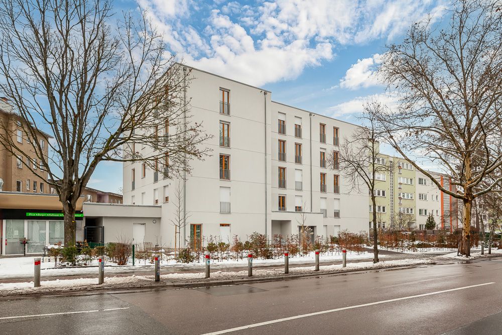 Modernes, vierstöckiges Wohnhaus mit vielen Fenstern, Bäumen und verschneitem Gehweg davor.