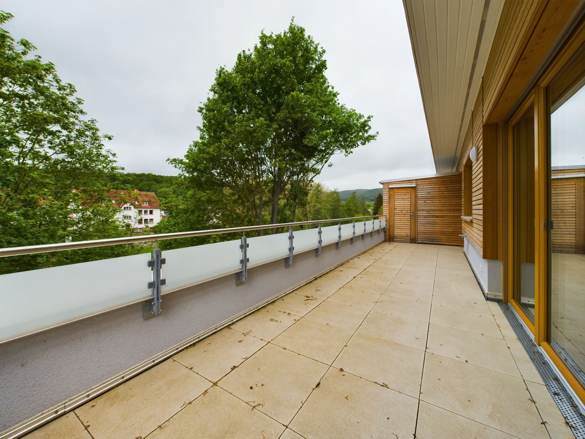 Großer Balkon mit Fliesenboden, Glasgeländer und Blick auf Bäume und benachbarte Häuser.