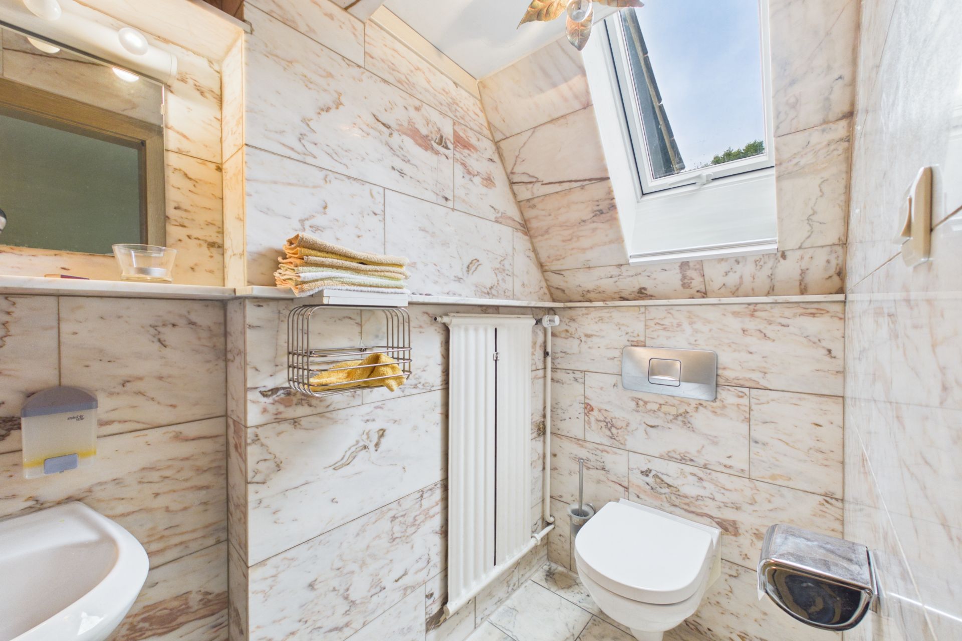 Kleines Badezimmer mit Marmorfliesen, Wandtoilette, Handtüchern und offenem Fenster.