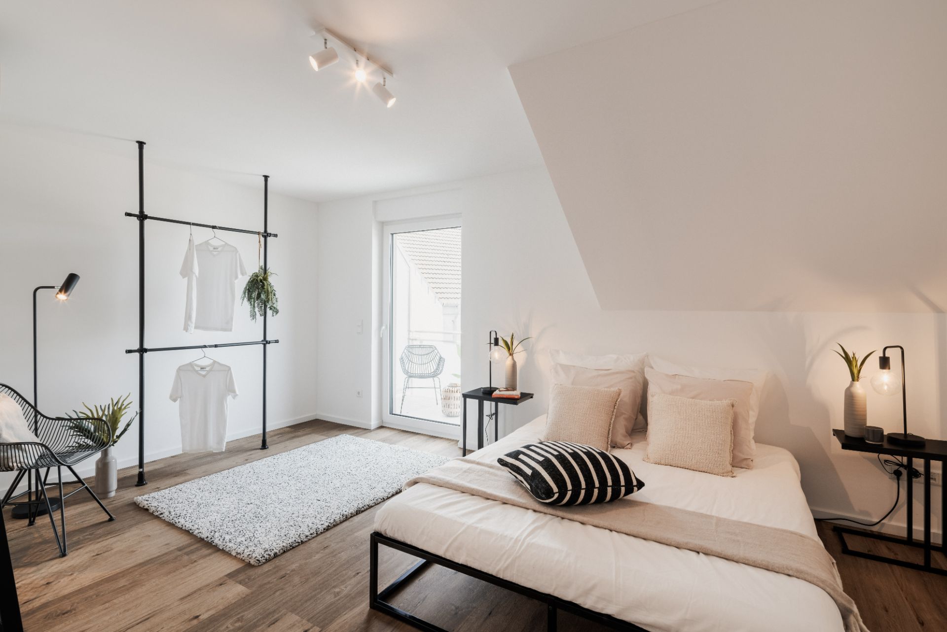 Helles Schlafzimmer mit Doppelbett, Nachttischen, Teppich, offener Kleiderstange und Zugang zum Balkon.