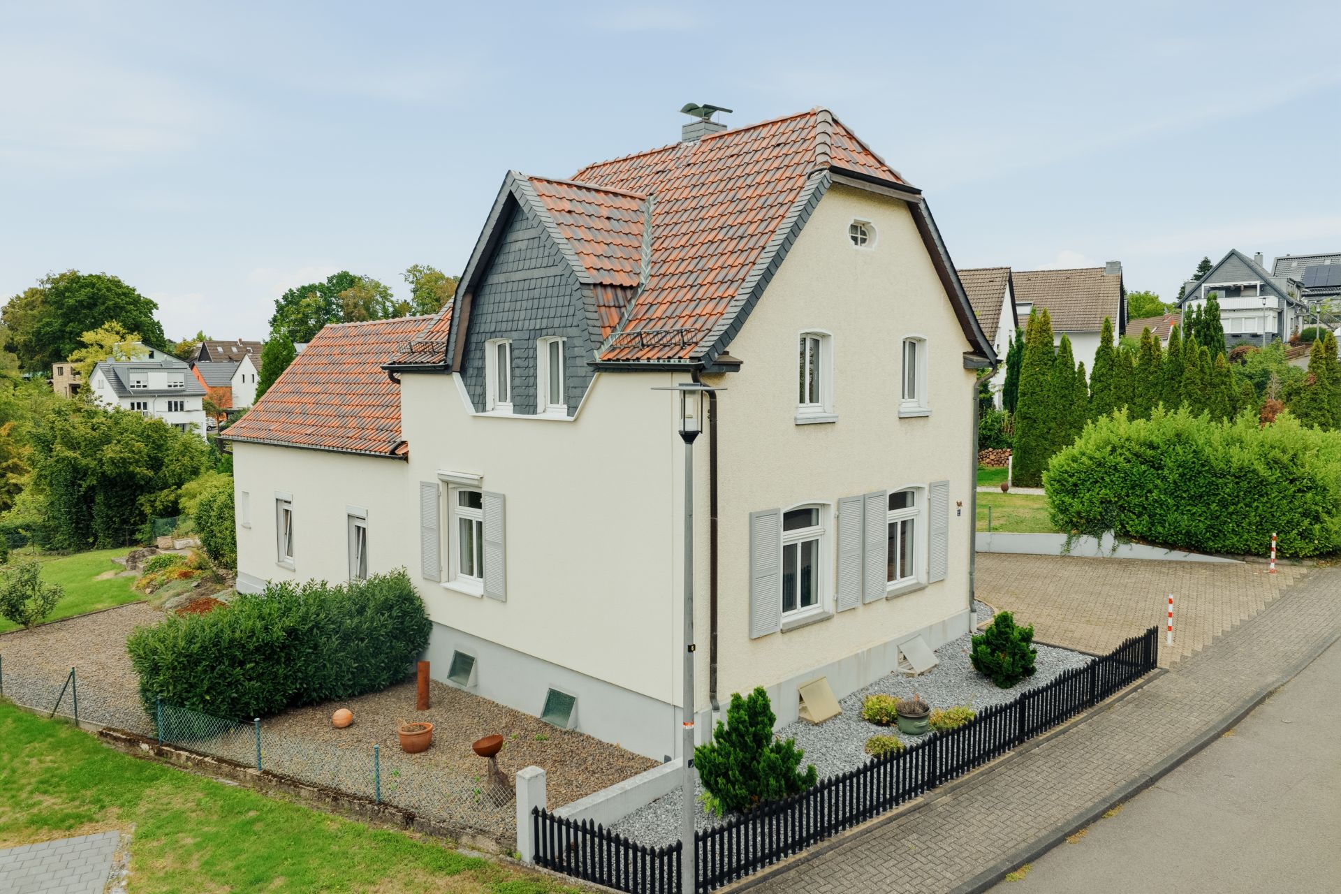 Zweistöckiges Haus mit grauem Zaun, rotem Dach und gepflegtem Garten an einer Straße.