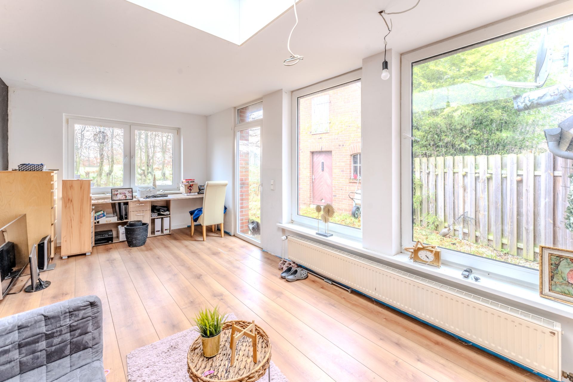 Helles Wohnzimmer mit großen Fenstern, Holzfußboden, Schreibtisch und Blick in den Garten.