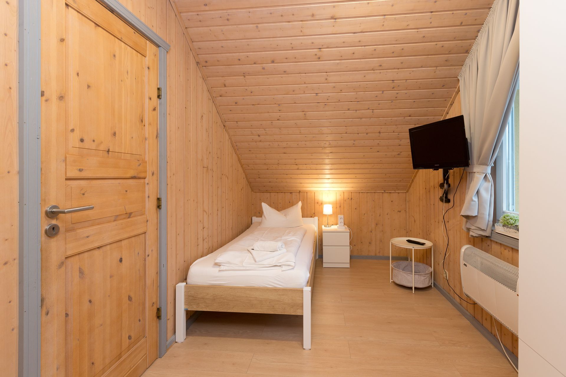 Kleines Schlafzimmer mit Einzelbett, Nachttisch, Lampe, TV und Holzverkleidung.