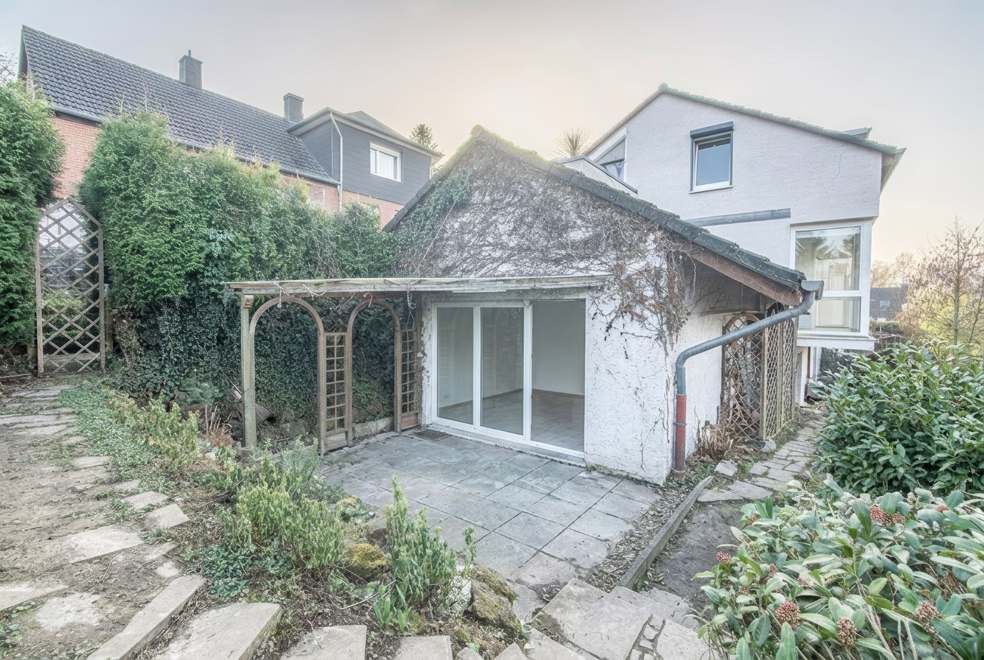 Kleines Gartenhaus mit großem Fenster, Terrasse und Rankgitter, umgeben von Pflanzen.