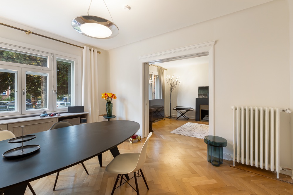 Esszimmer mit ovalem Tisch, weißen Stühlen, Holzfußboden und Blick ins angrenzende Wohnzimmer.