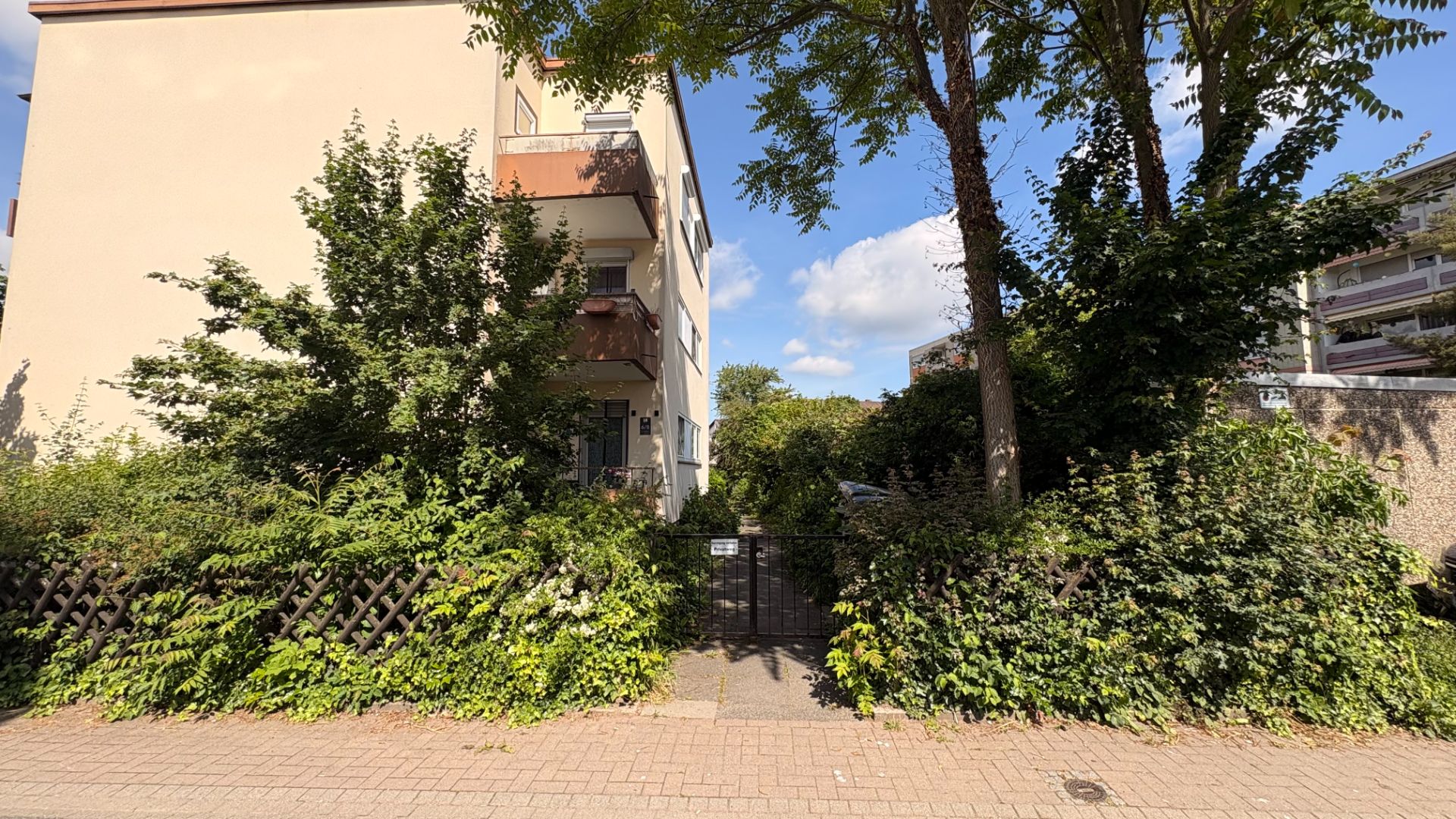Eingang zu einem Wohnhaus mit Garten und Bäumen, umgeben von Hecken und Zaun.