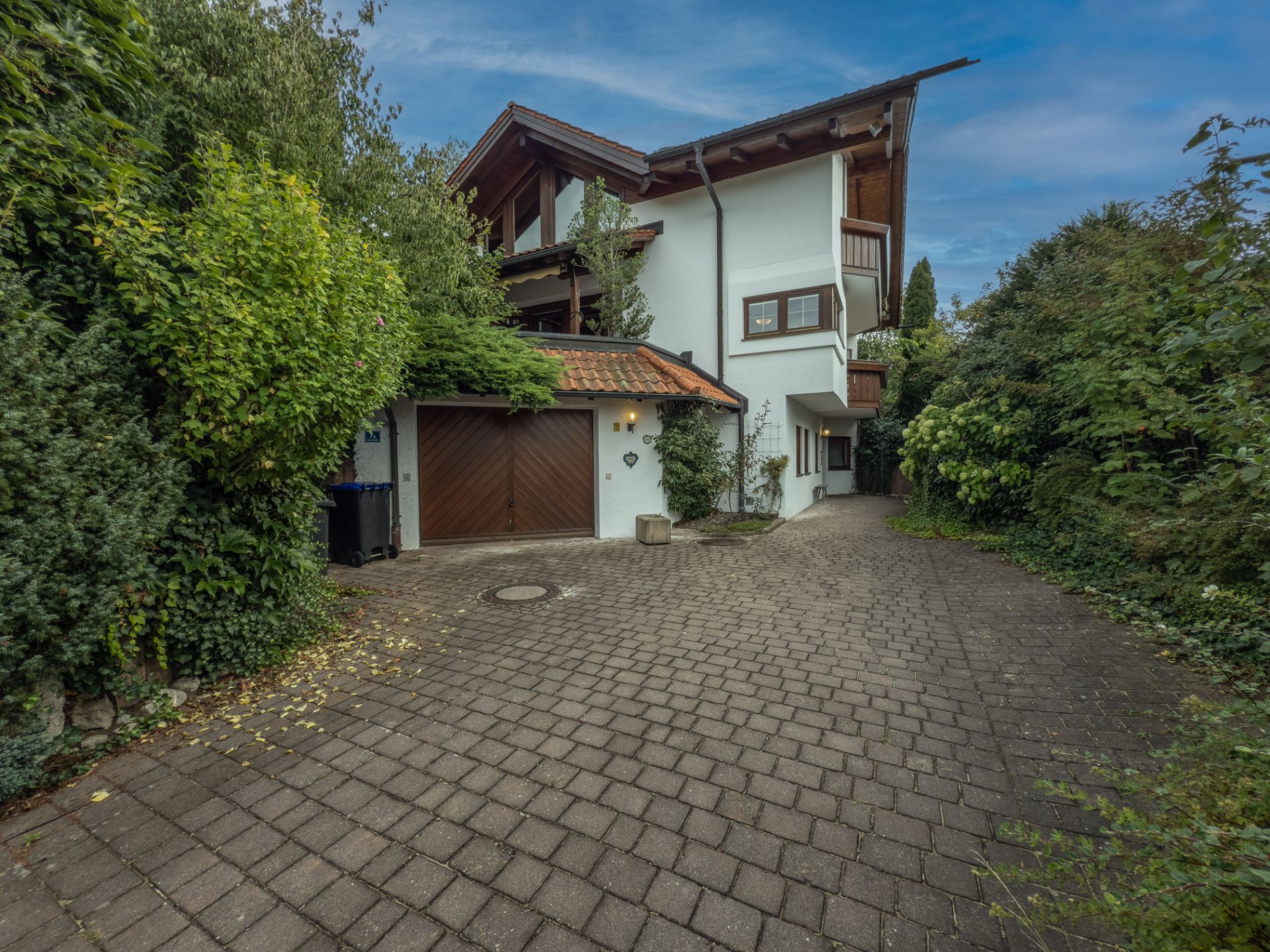 Einfamilienhaus mit weißer Fassade, Doppelgarage und gepflasterter Einfahrt, umgeben von viel Grün.
