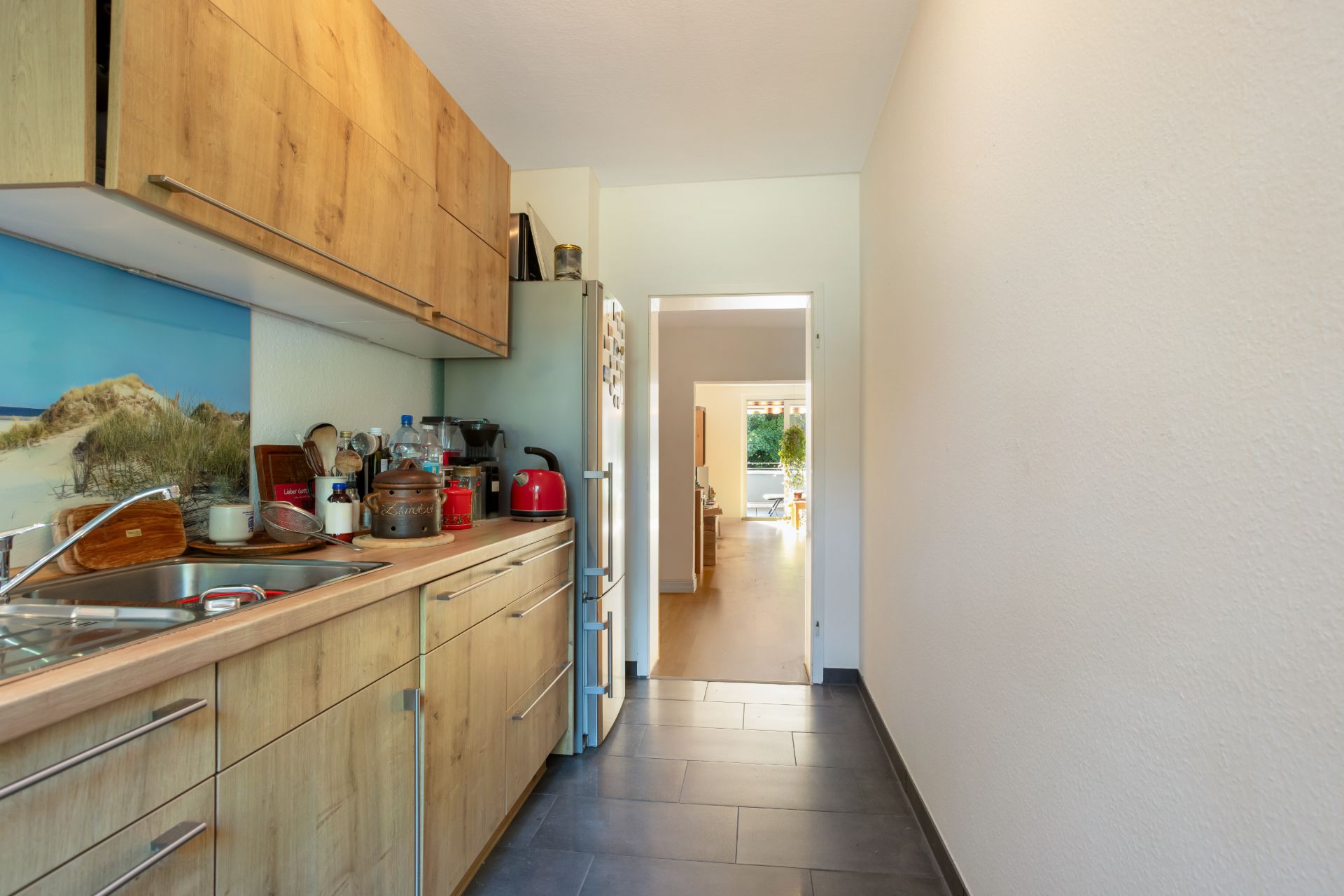 Schmale Küche mit Holzmöbeln, Spüle, Kühlschrank und Geräten, Tür zum hellen Wohnzimmer.