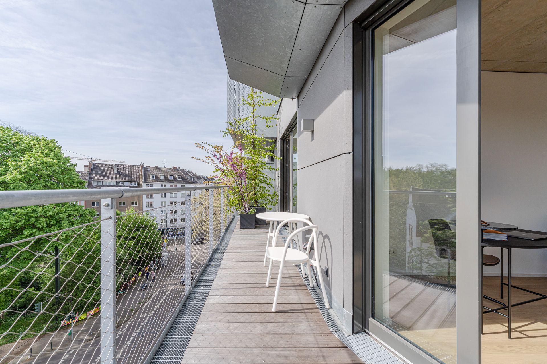 Moderner Balkon mit zwei weißen Stühlen, Tisch und Pflanze, Blick auf Straße und Bäume.