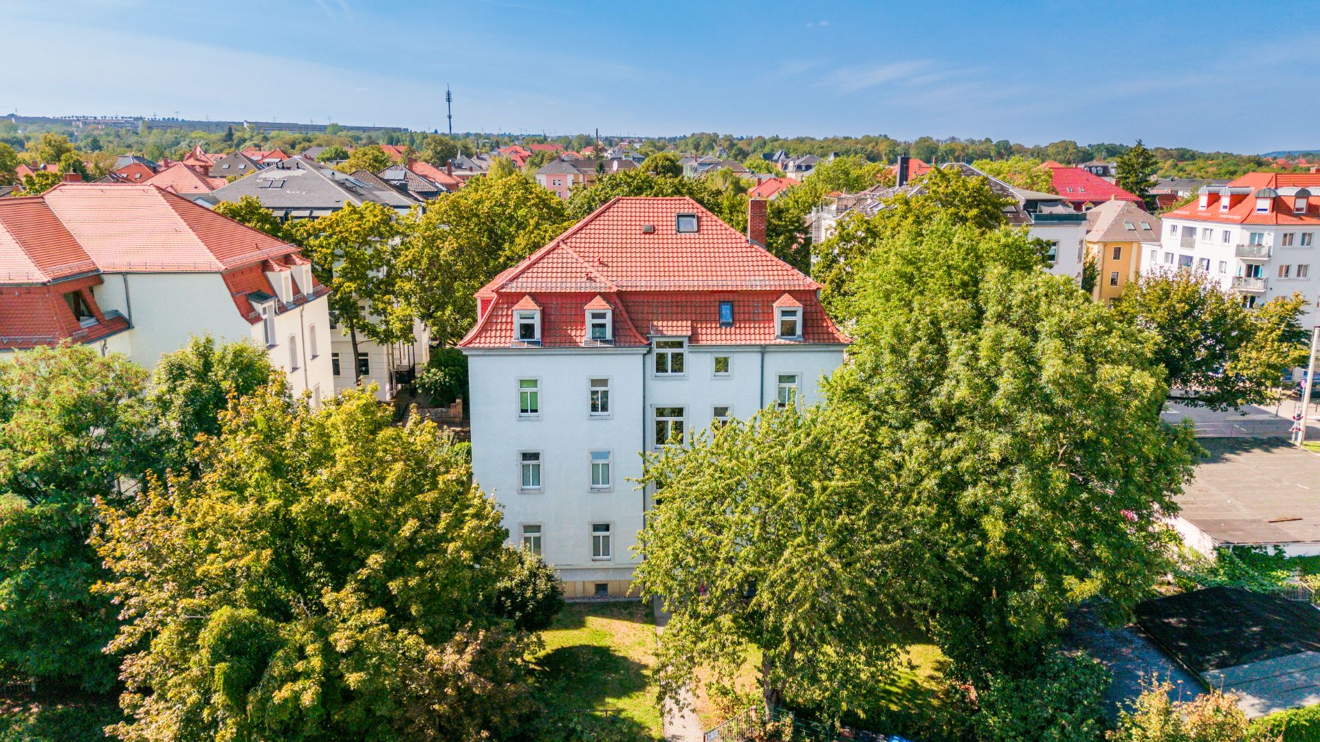 Mehrstöckiges Wohnhaus mit rotem Dach, umgeben von Bäumen und Nachbargebäuden.