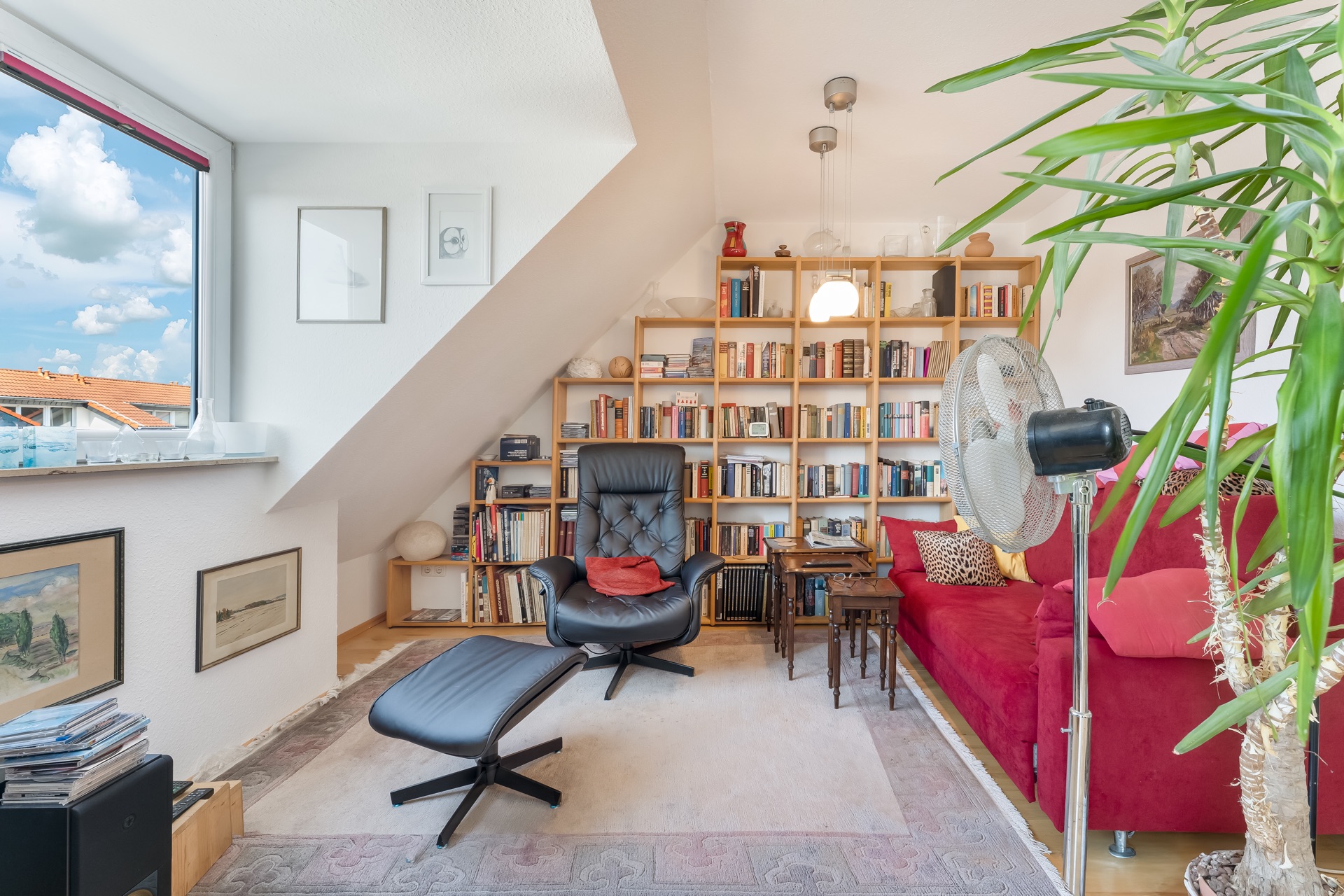Wohnzimmer mit rotem Sofa, Sessel, Bücherregal, großem Fenster und Zimmerpflanze.
