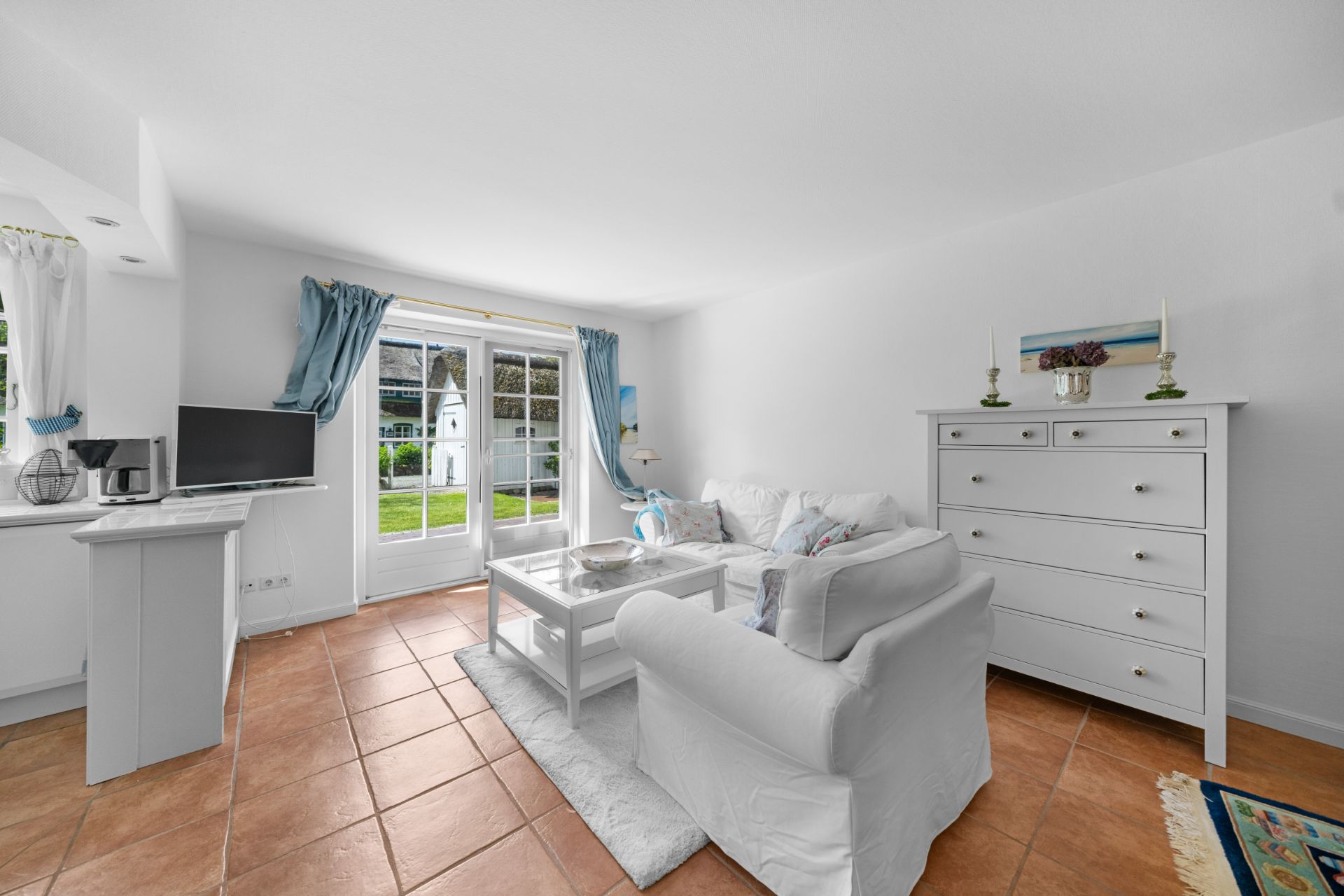 Helles Wohnzimmer mit weißem Sofa, Kommode und Zugang zum Garten durch Glastüren.