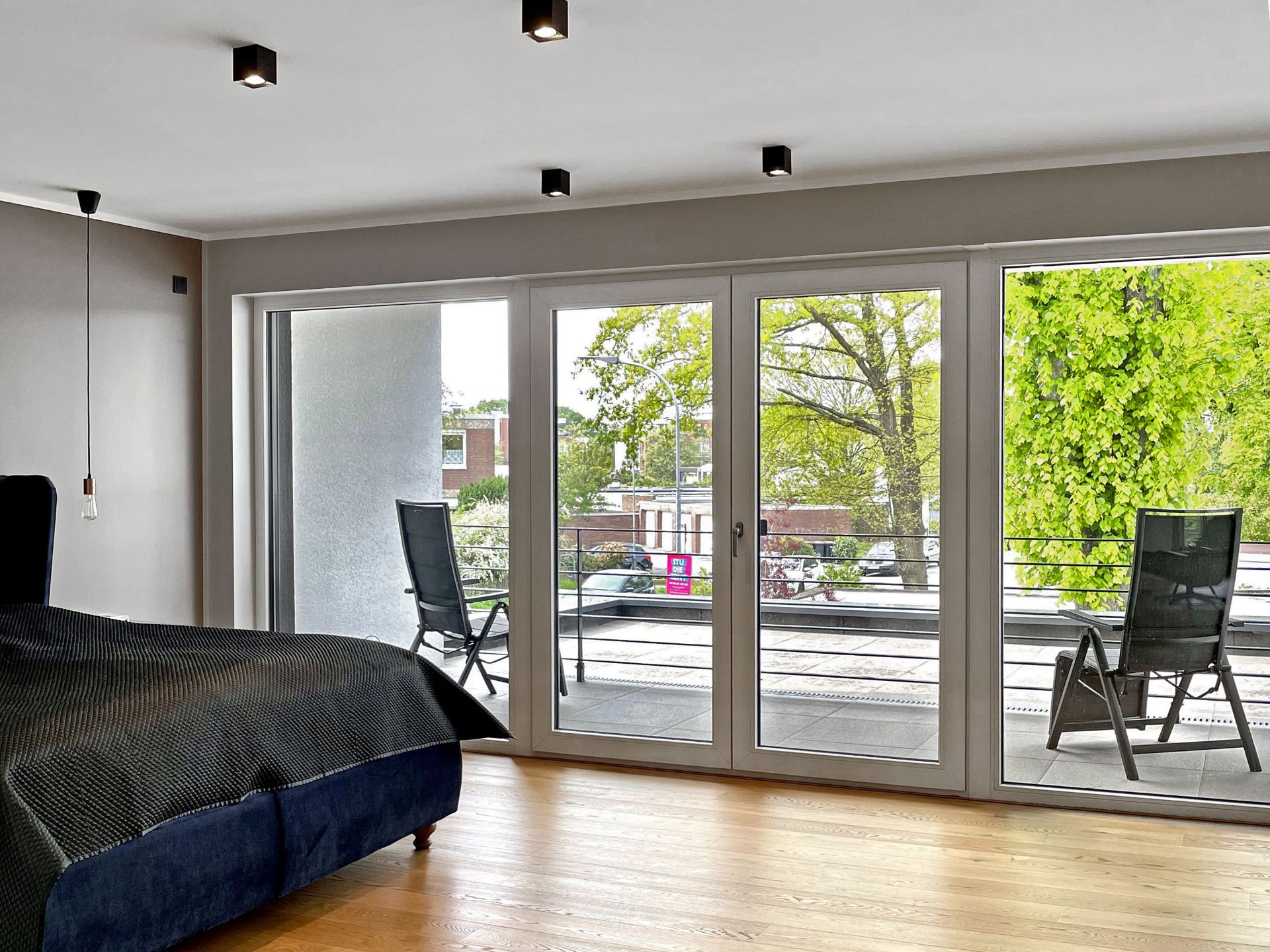 Schlafzimmer mit Glasfront und Zugang zu Balkon mit zwei Stühlen, Blick auf Bäume.