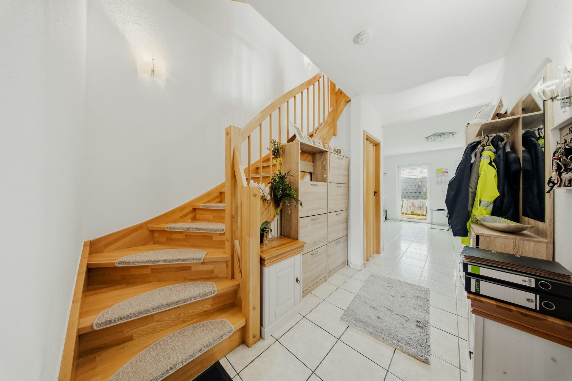 Helle Diele mit Holztreppe, Fliesenboden, Garderobe und Blick in einen weiteren Raum.