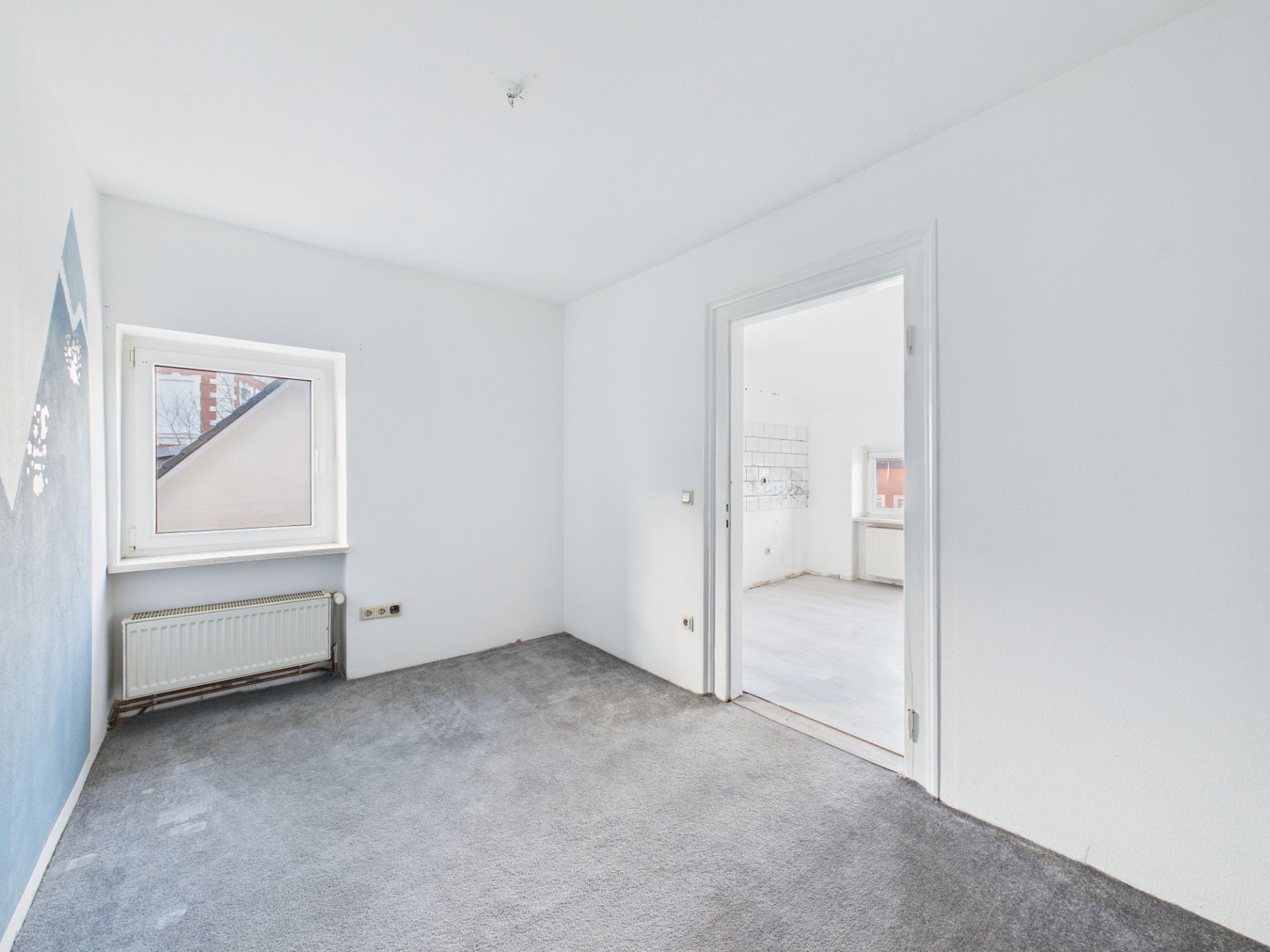 Helles Zimmer mit grauem Teppich, weißer Wand, Fenster und offener Tür zu weiterem Raum.