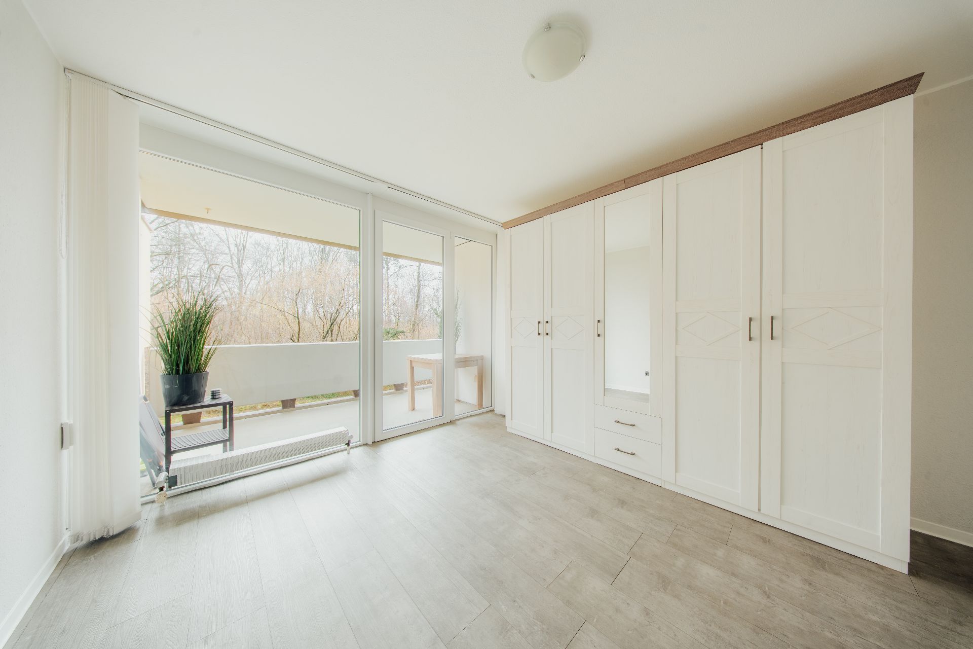 Helles Zimmer mit großem weißen Kleiderschrank und Zugang zum Balkon mit Pflanzen.