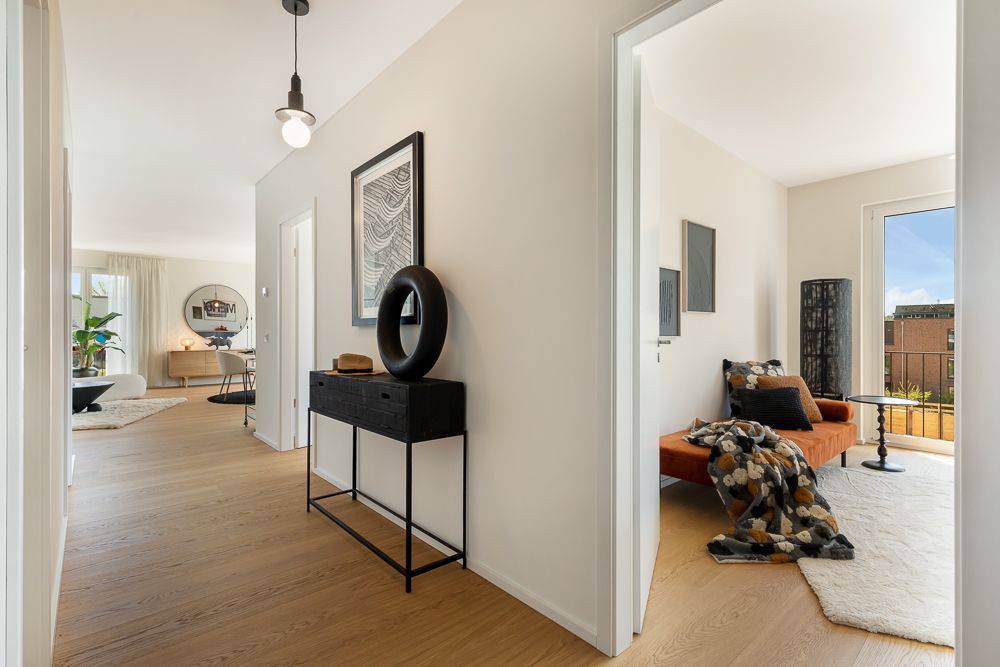 Heller Flur mit Holzfußboden, links Wohnbereich, rechts Zimmer mit Tageslicht und orangefarbener Couch.