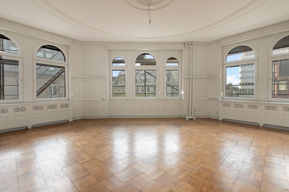 Großer, leerer Raum mit Holzboden und mehreren Fenstern, die auf eine Stadt zeigen.