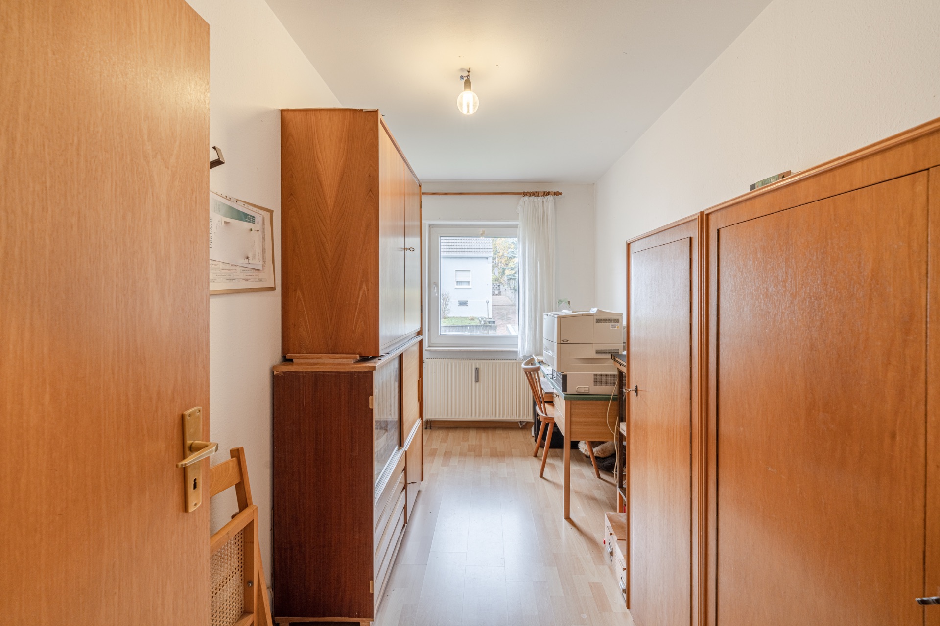 Kleines, schmales Zimmer mit Holzschränken, Schreibtisch und Fenster am Ende.