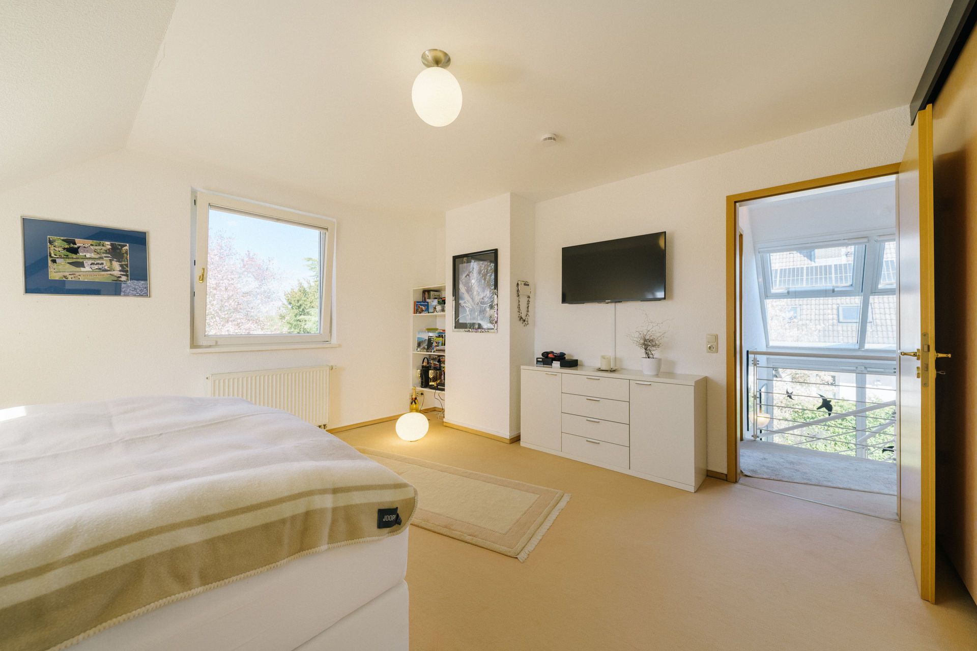 Helles Schlafzimmer mit Bett, Teppich, Kommode, Wandfernseher und Fenster mit Blick nach draußen.
