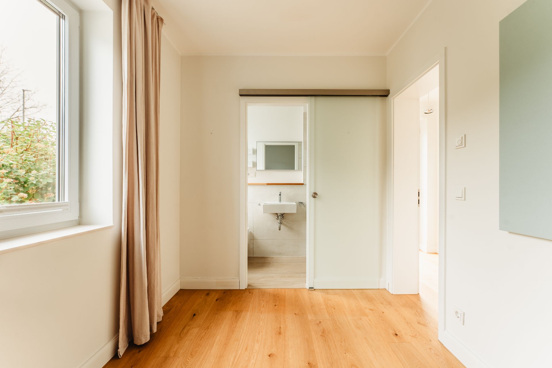 Helles Zimmer mit Holzfußboden, großem Fenster, beigen Vorhängen und Blick ins Badezimmer mit Schiebetür.