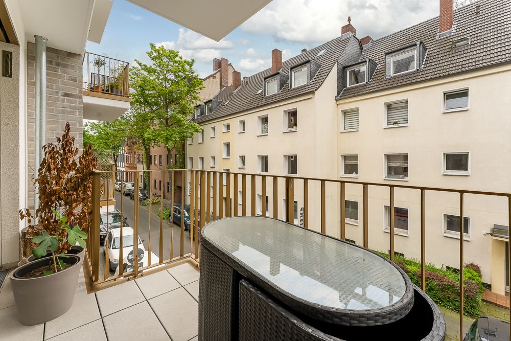 Balkon mit Glastisch, zwei Stühlen, Pflanzen und Blick auf eine Wohnstraße mit Mehrfamilienhäusern.
