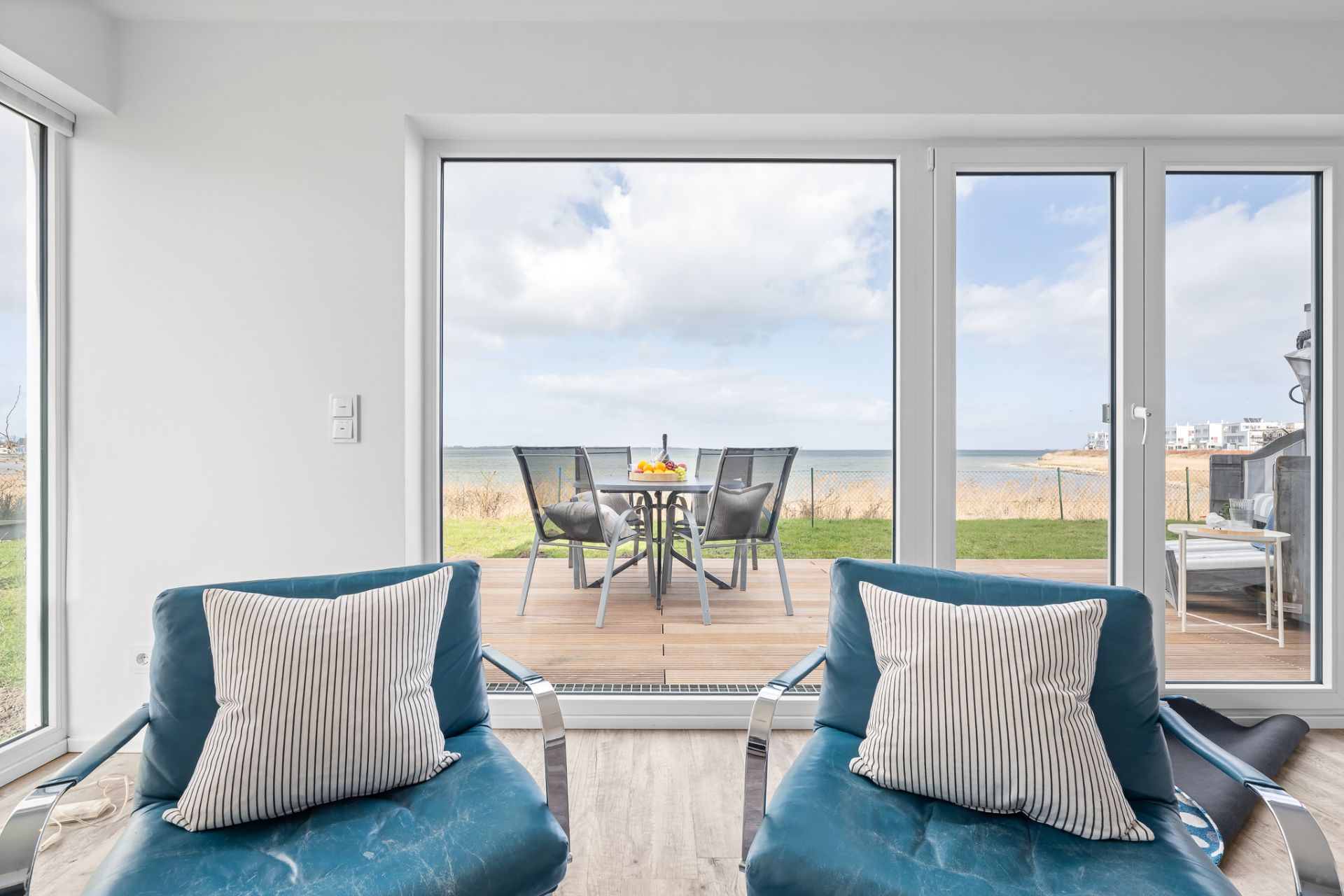Zwei Sessel mit Kissen vor großen Fenstern, dahinter Terrasse mit Tisch und Meerblick.