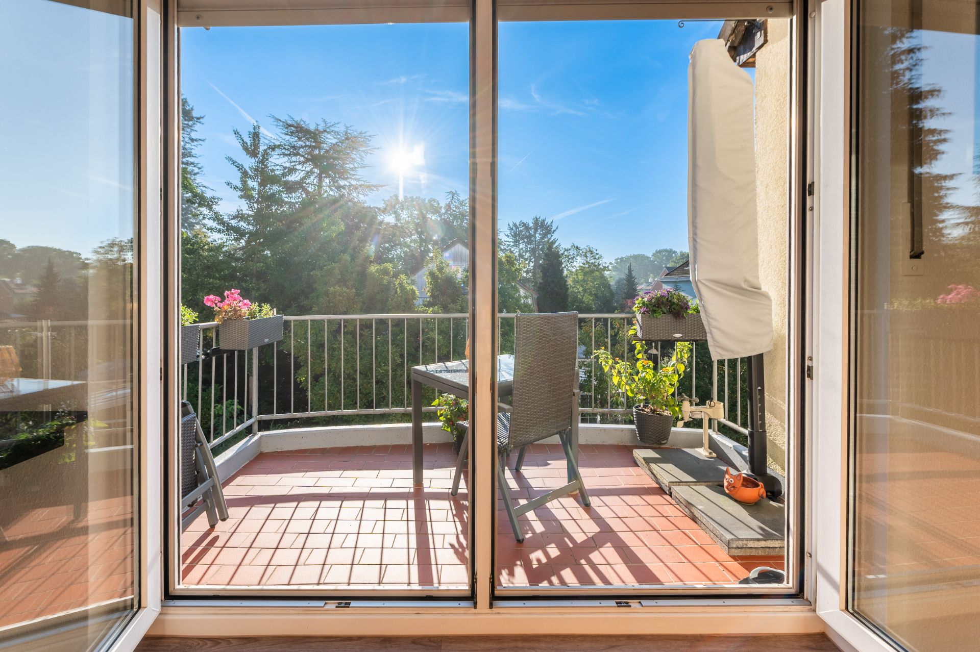 Balkon mit Tisch, Stühlen und Pflanzen, Blick durch eine offene Glastür, sonniges Wetter.