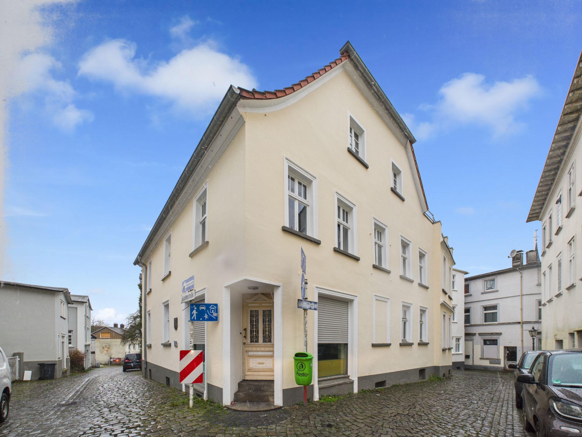 Ecke eines hellen Wohnhauses an einer gepflasterten Straße mit mehreren Fenstern und Eingangstür.