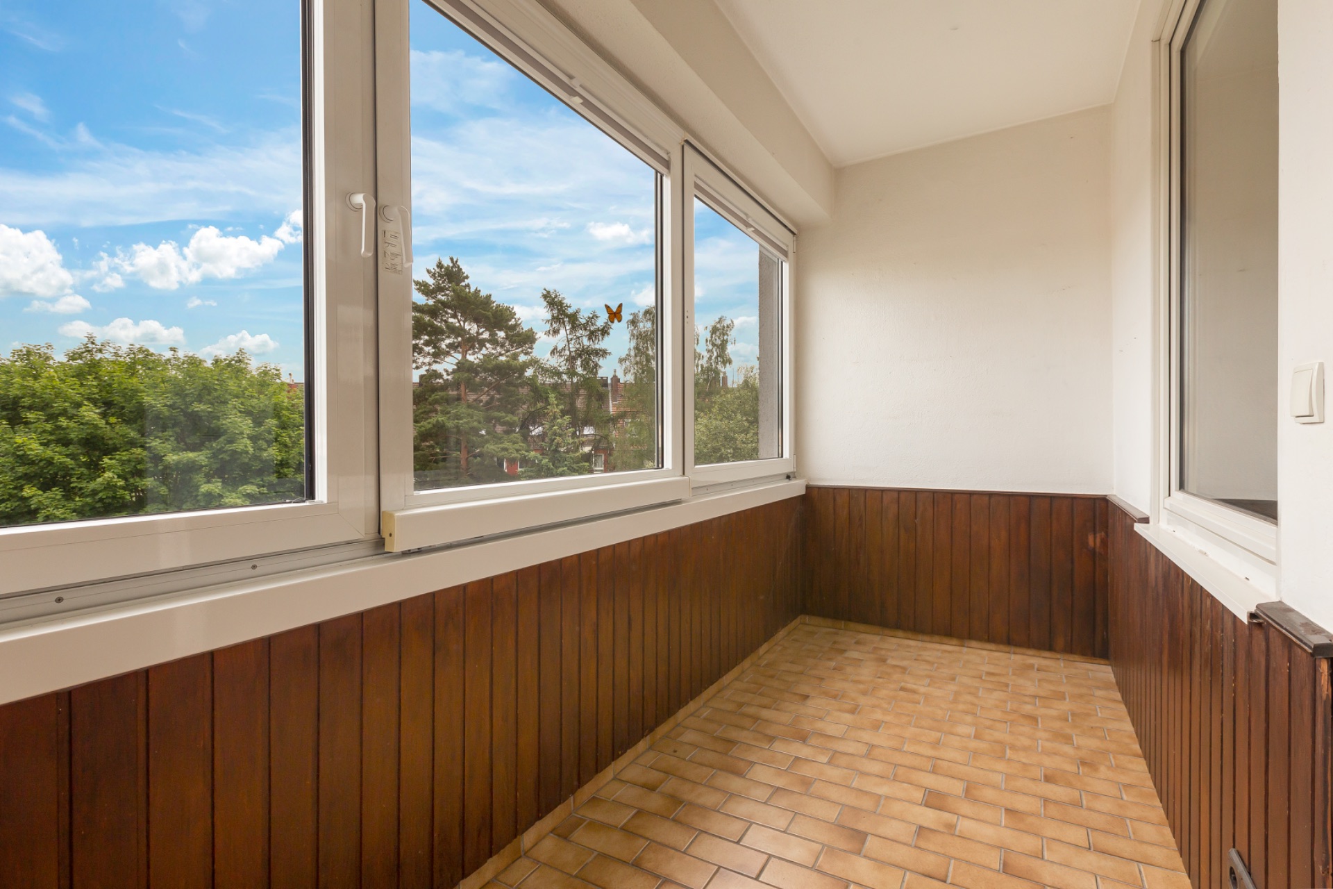 Verglaster Balkon mit Holzwandverkleidung und Fliesenboden, Blick ins Grüne.