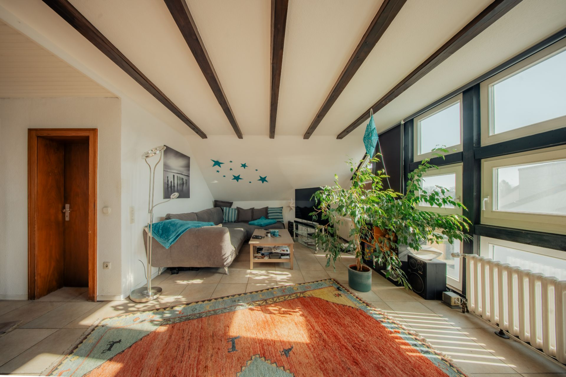 Helles Wohnzimmer mit Sofa, großem Teppich, Zimmerpflanze und großen Fenstern.