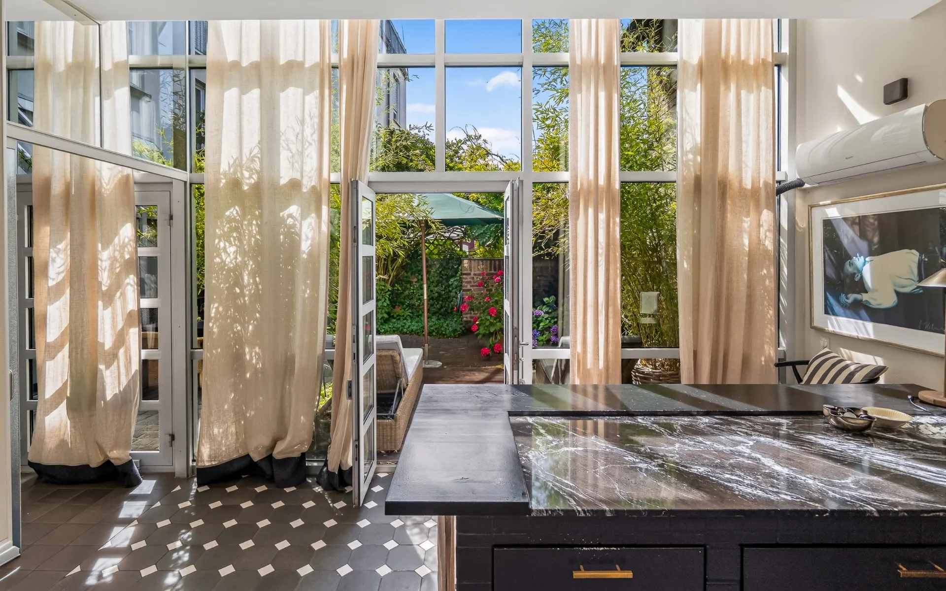 Helles Wohnzimmer mit großen Fenstern, Vorhängen und Blick auf einen Garten mit Pflanzen und Sonnenschirm.