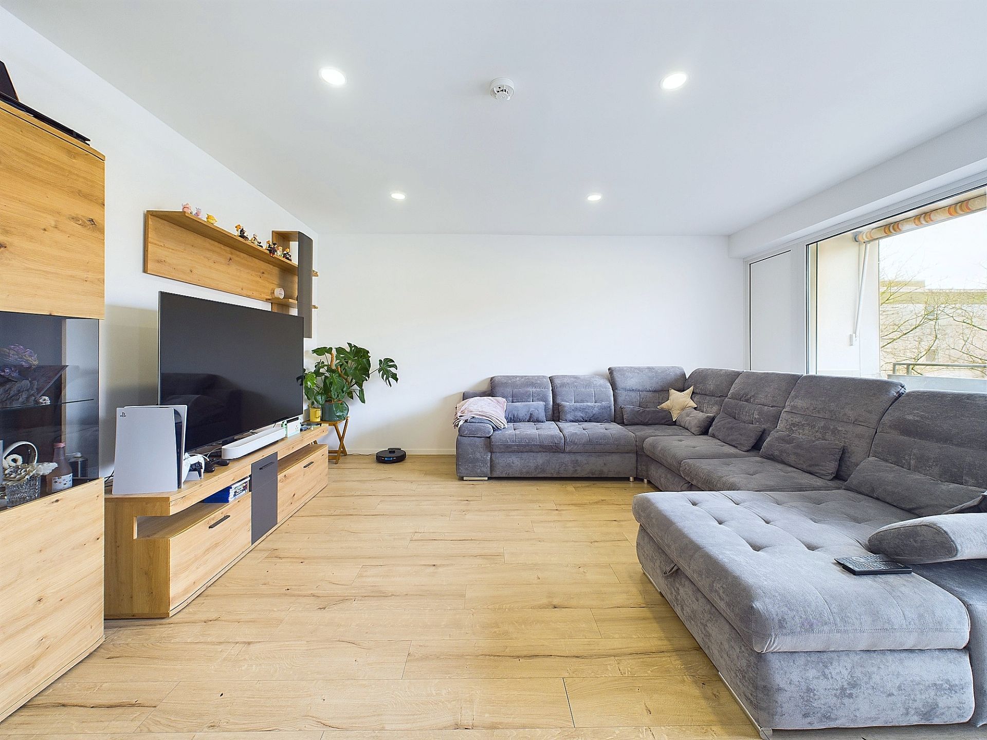 Helles Wohnzimmer mit grauem Ecksofa, großem Fernseher und Holzregalen auf hellem Holzboden.