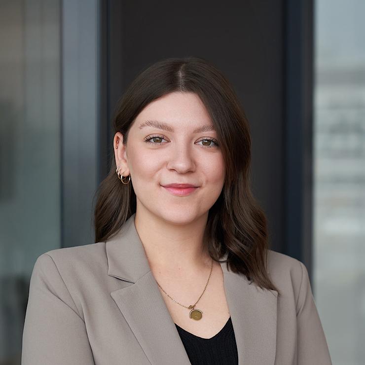Junge Frau mit braunen Haaren im beigen Blazer steht vor einem Glasfenster.
