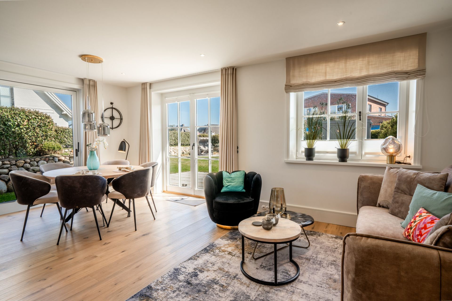 Helles Wohnzimmer mit Sofa, Sessel, Couchtischen, Esstisch und großen Fenstern zum Garten.