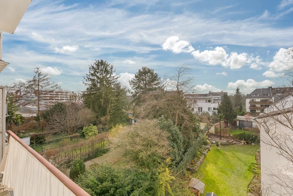 Blick vom Balkon auf einen Garten mit Bäumen und Rasen, umgeben von Wohnhäusern.