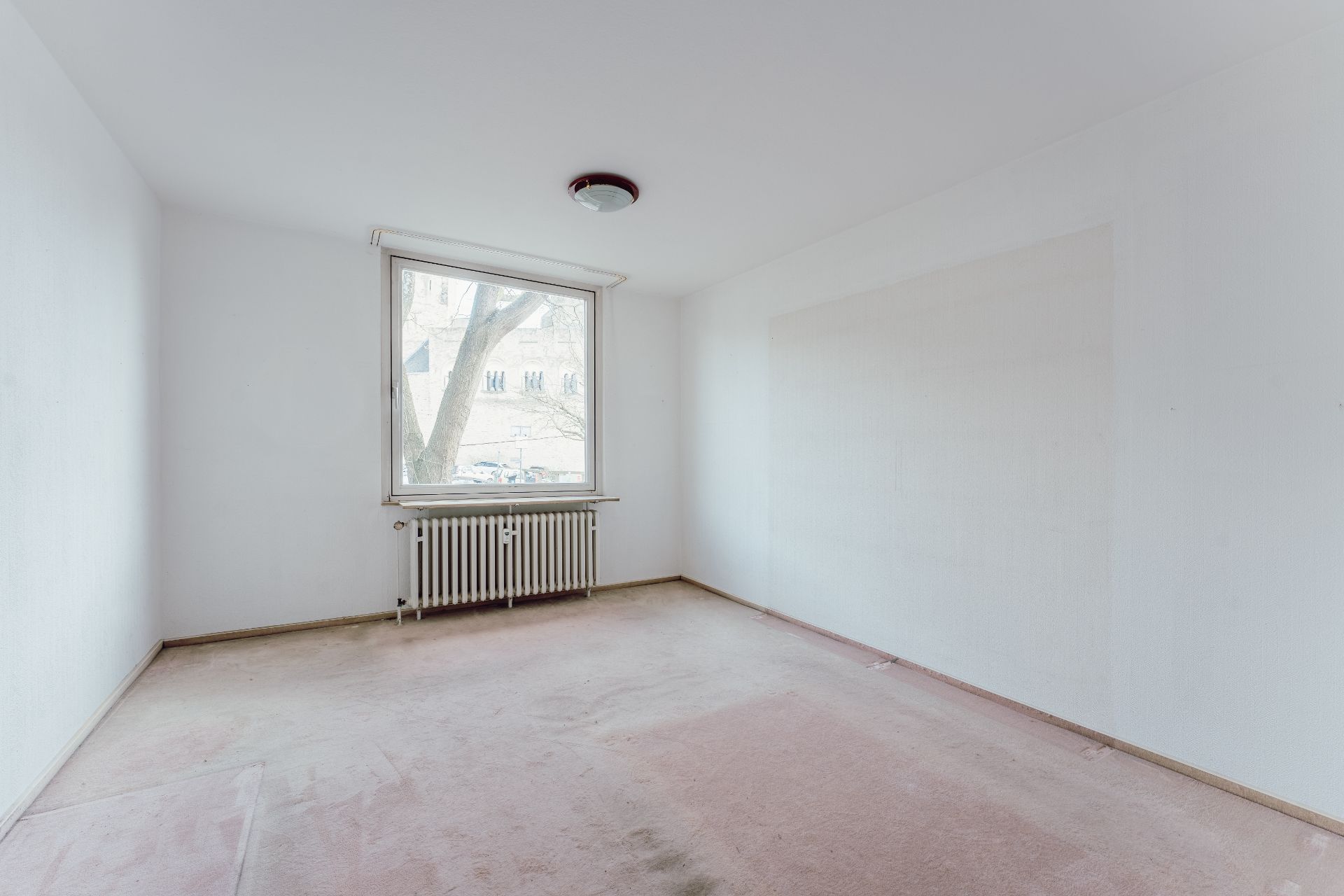 Leeres Zimmer mit großem Fenster, Heizkörper und hellem Teppichboden.