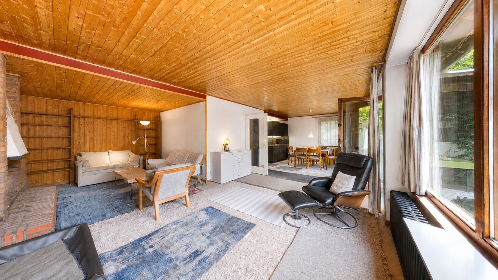 Helles Wohnzimmer mit Holzboden, Teppichen, Sesseln, Sofa und großem Fenster mit Blick ins Grüne.